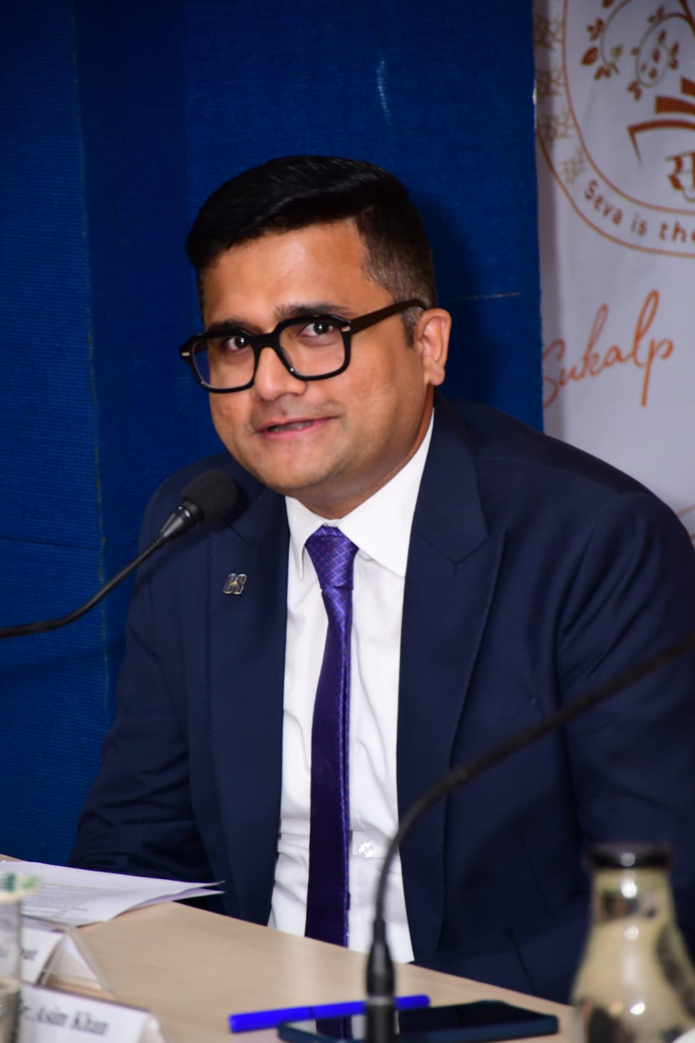 Candidate in navy blue suit with purple tie speaking at a conference panel setting